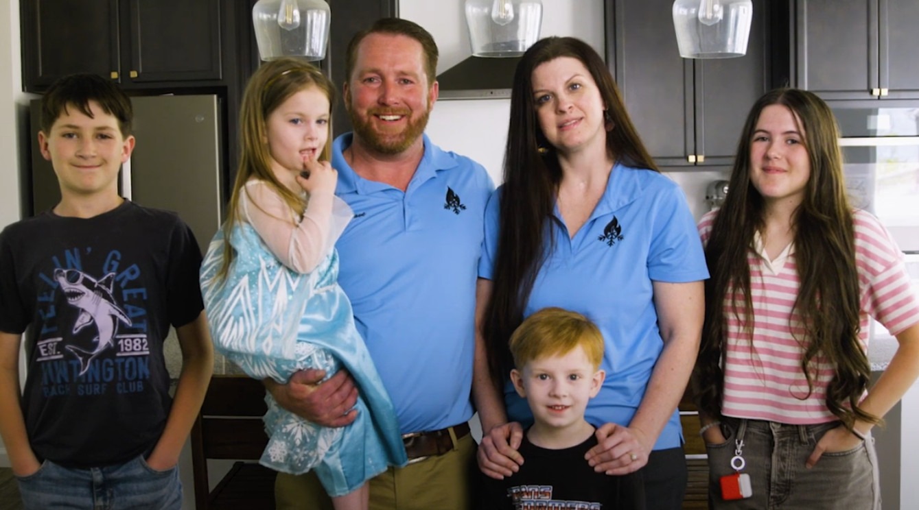 A smiling family of six standing together in a modern kitchen, with two adults wearing Proformance HVAC uniforms and four children gathered around them, representing the team behind the local Utah HVAC company.